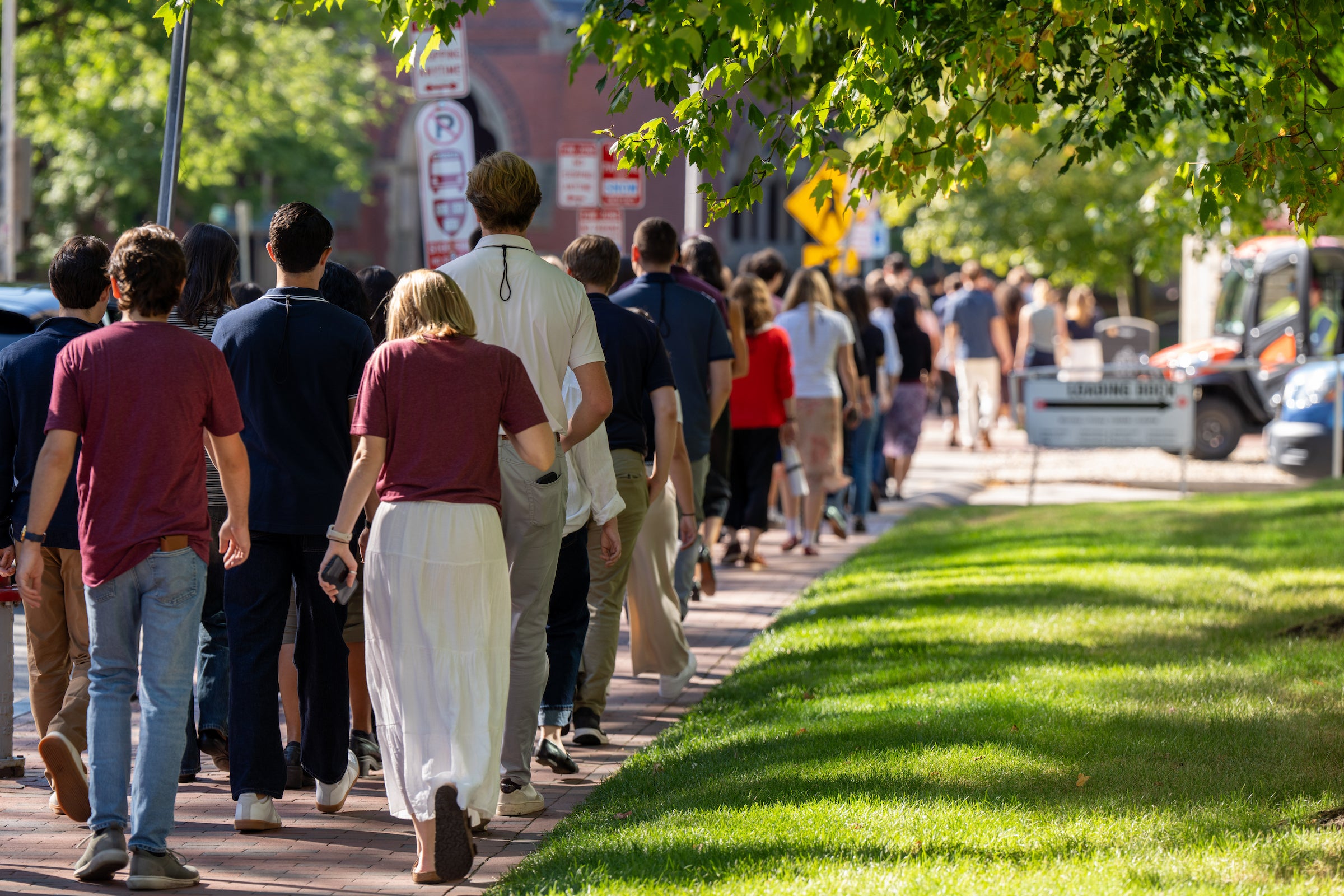 A group of students walks away from the camera along a sunlit paved path lined with trees and street signs.