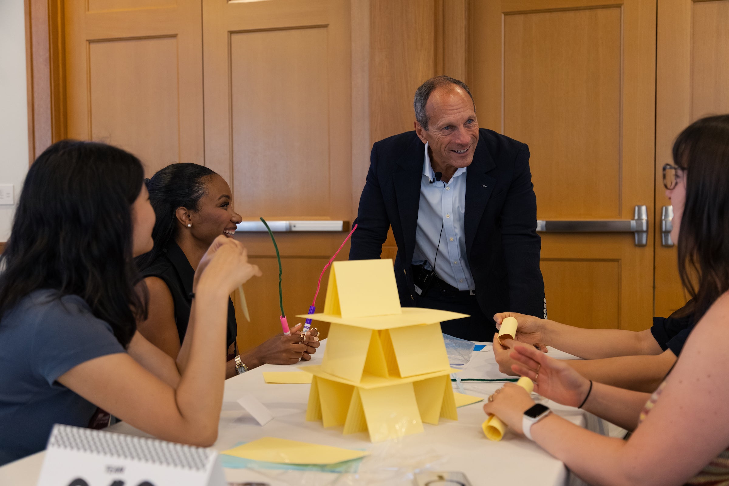 A group of students collaboratively build a structure from yellow paper at a table, while a professor leans over the table and converses with them.