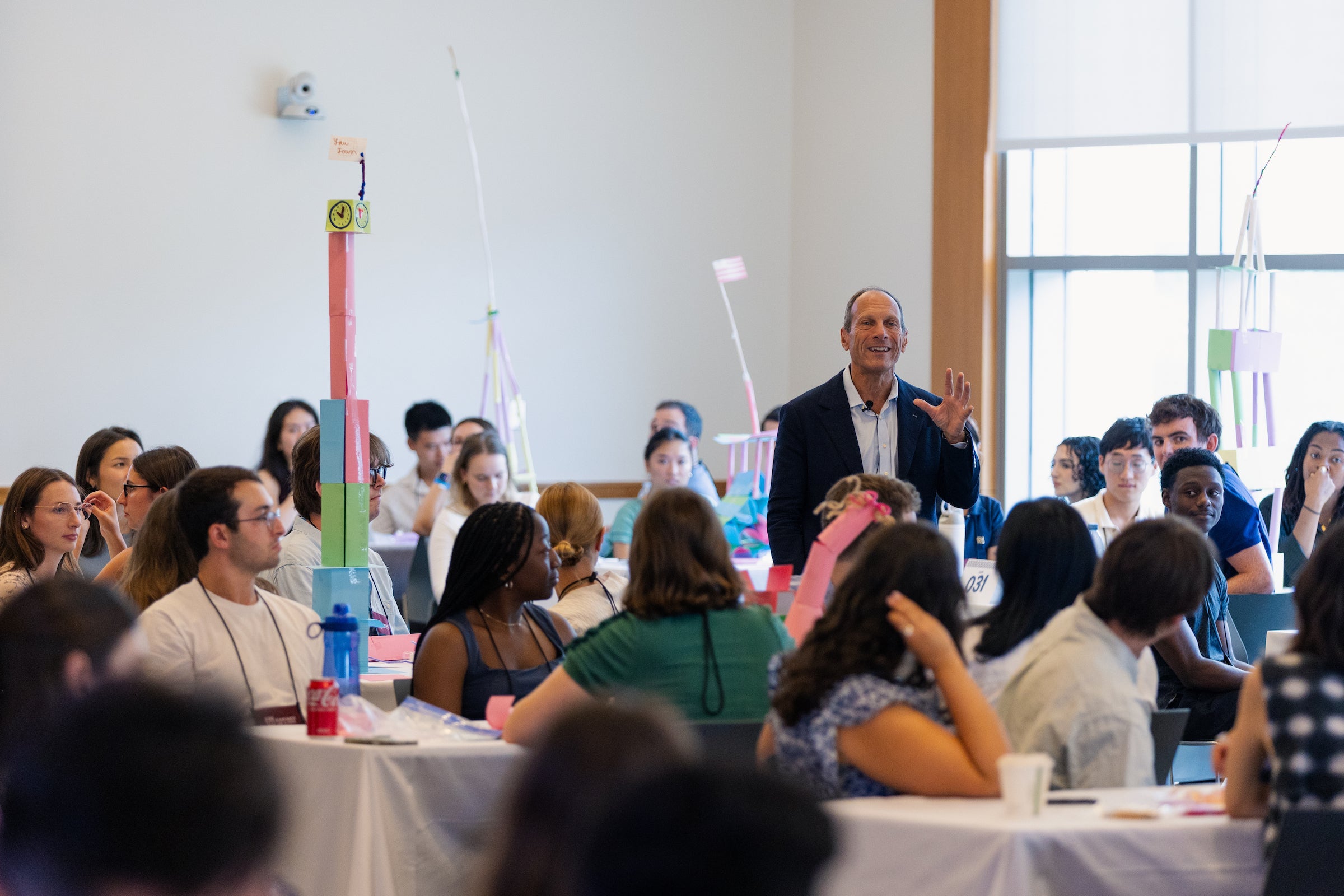 A speaker addresses an audience seated at tables in a brightly lit room, with colorful handmade structures visible in the background.