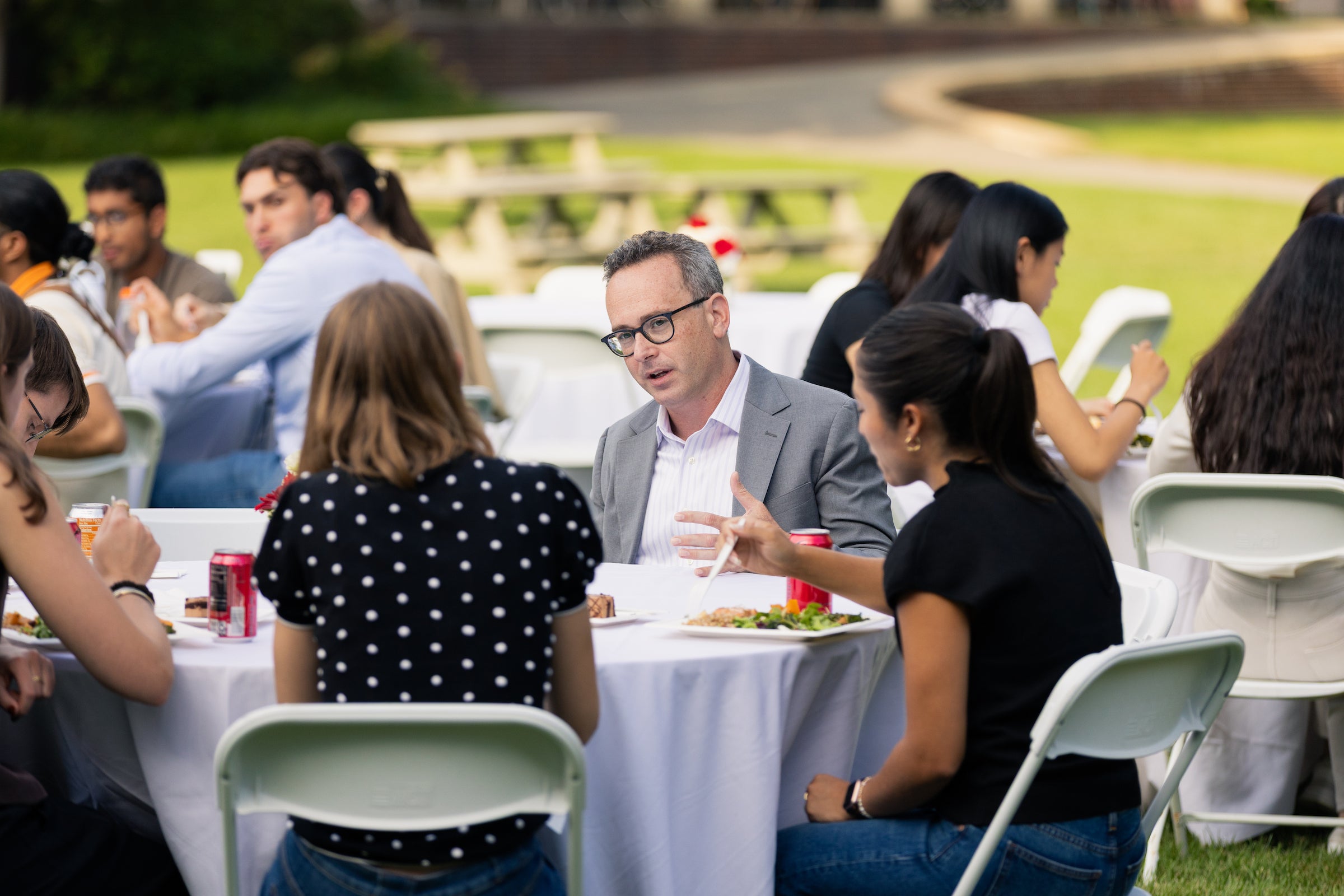 A professor converses with students while seated at a round table at an outdoor event.