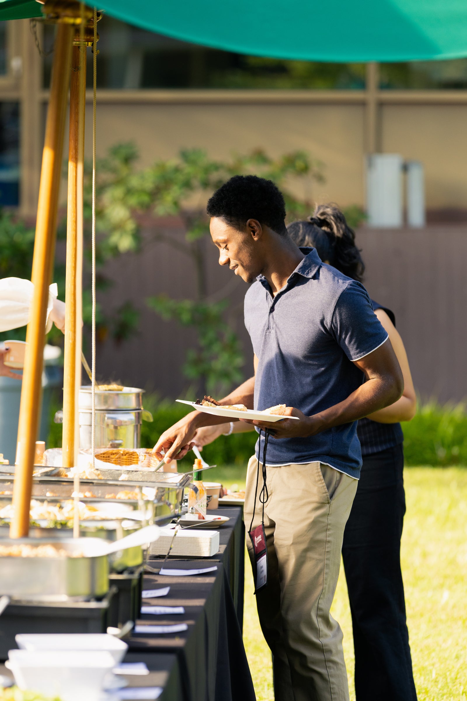 A student selects food from a buffet under a green umbrella at a sunny outdoor event.