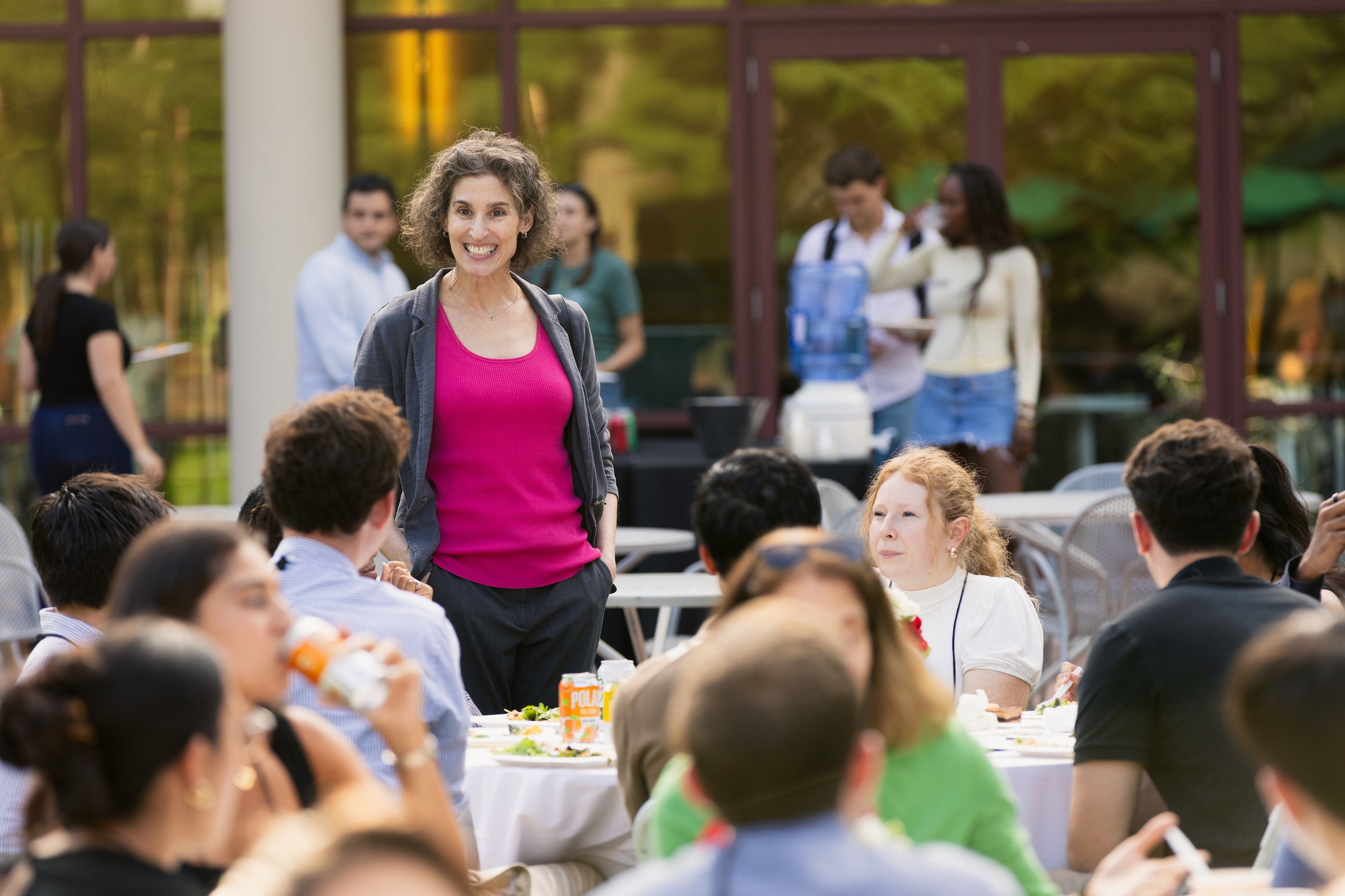 A professor converses with students seated at an outdoor dining table during a social gathering.