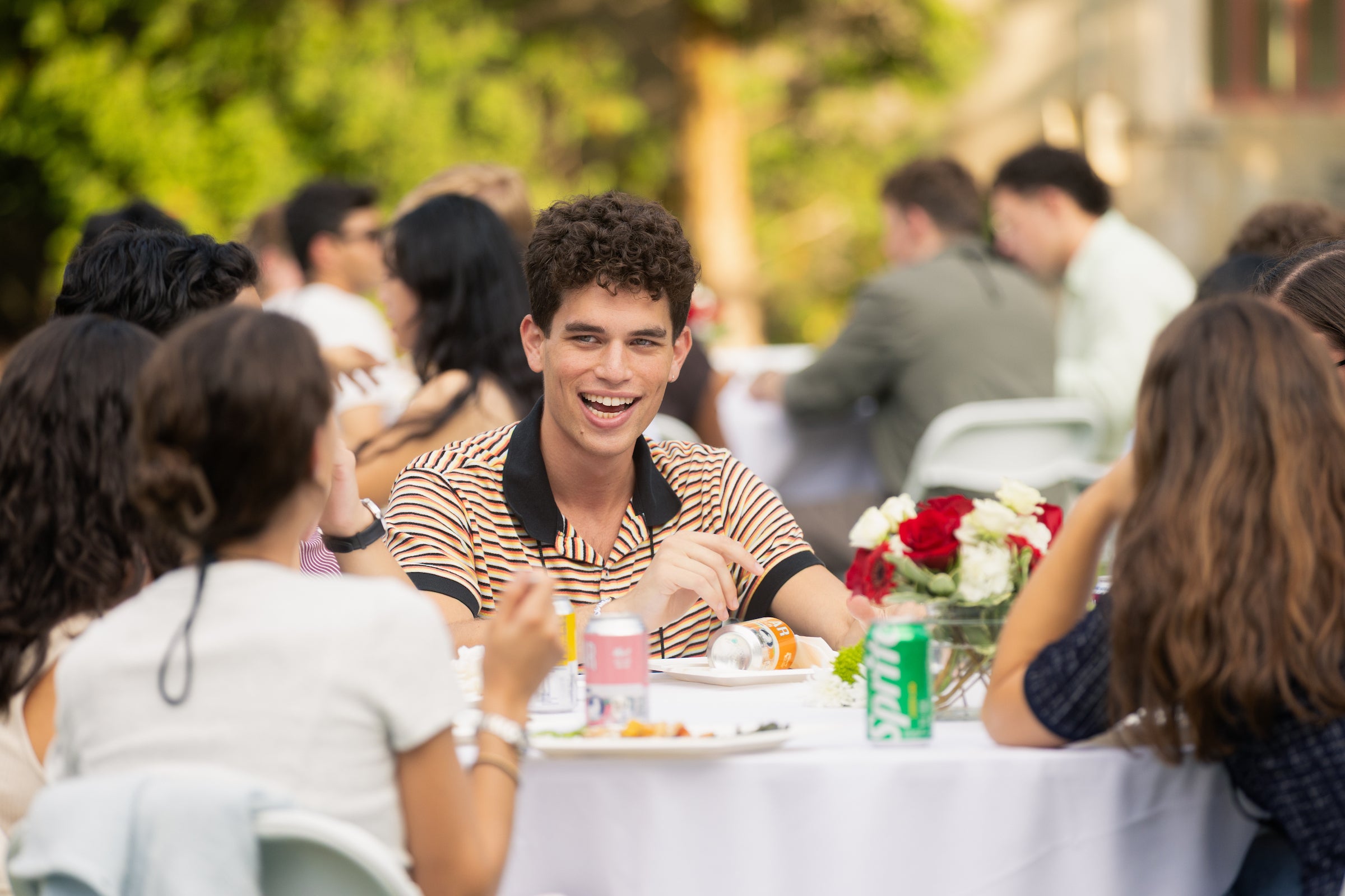 Students laugh and converse while seated at an outdoor dining table surrounded by others during a social gathering.