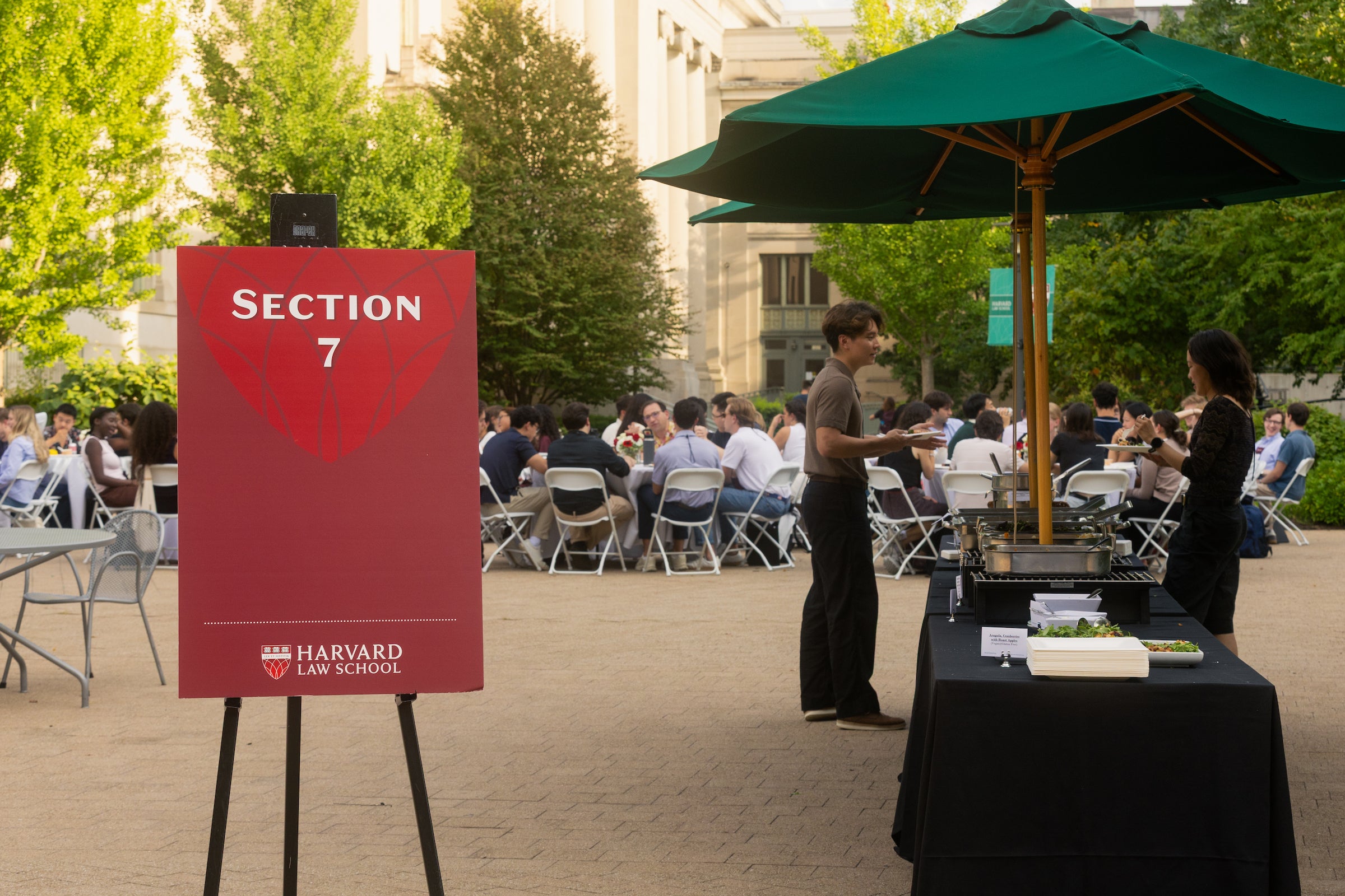 A red sign for Section 7 stands outdoors near a food station under a green umbrella, with people seated at tables in the background.