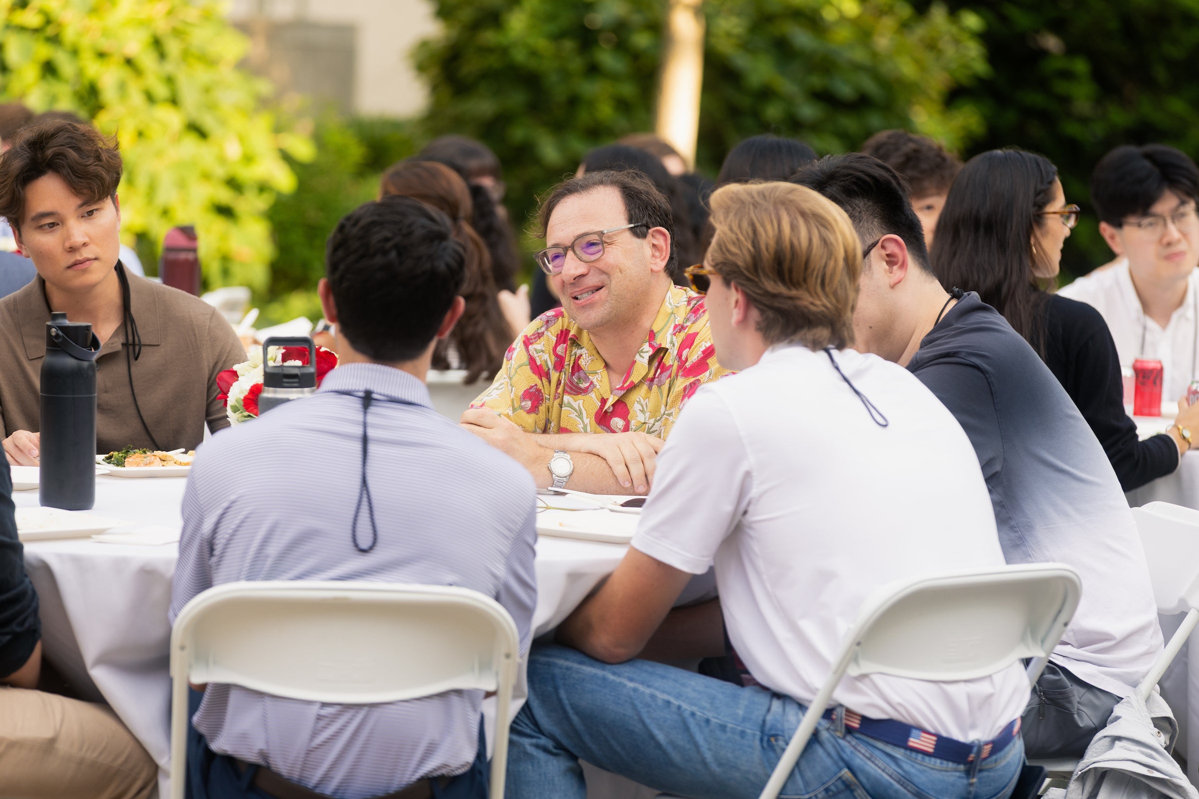 A professor converses with students while seated at a round table at an outdoor event.