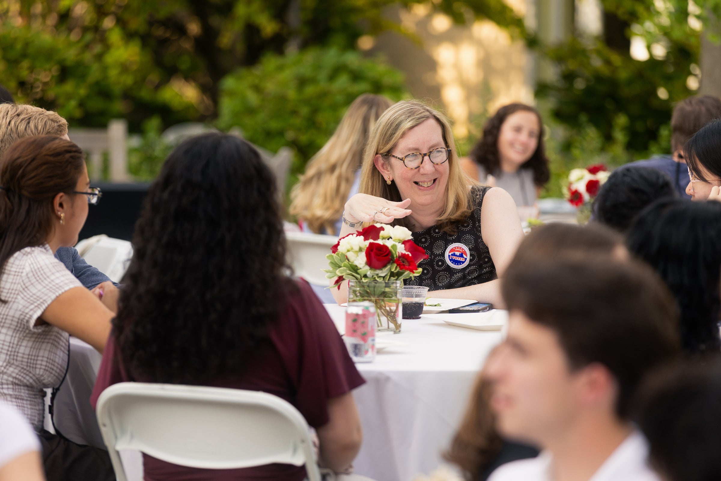 A professor converses with students while seated at a round table at an outdoor event.