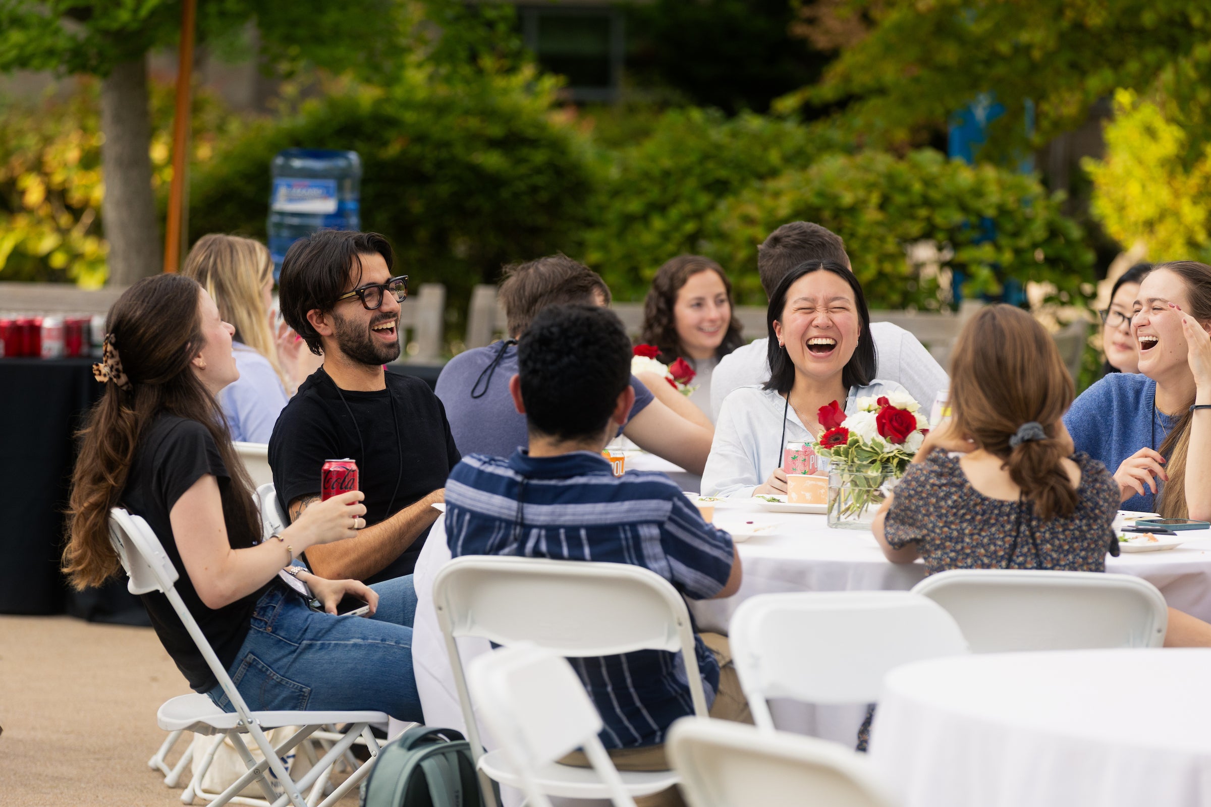 A group of students are laughing and socializing outdoors at a table set with drinks and flowers.