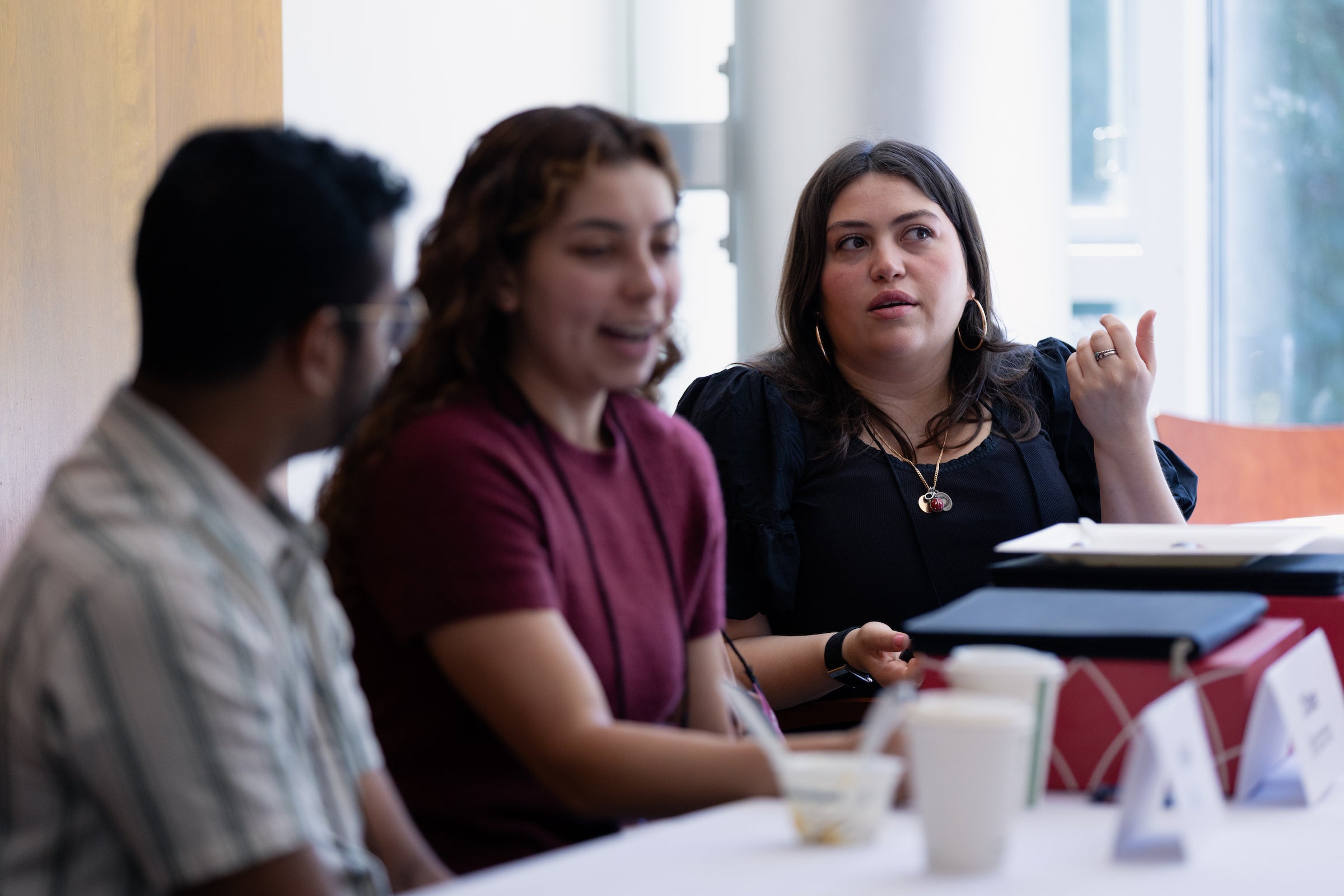 Three students engage in conversation around a table in a brightly lit room.