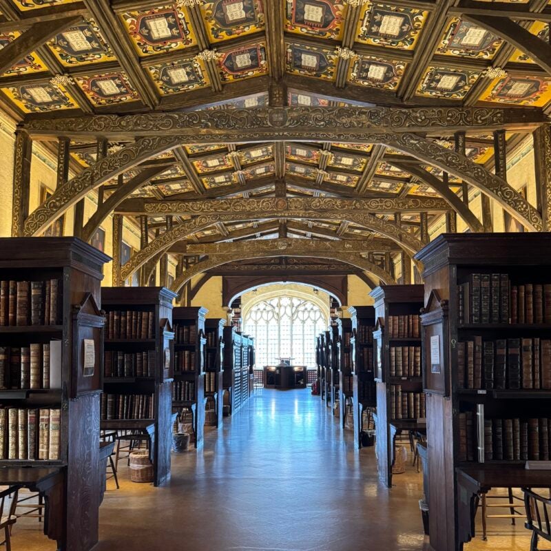 Long view of a library with ornate vaulted ceiling, bookcases on both sides, and a bright window in the distance.