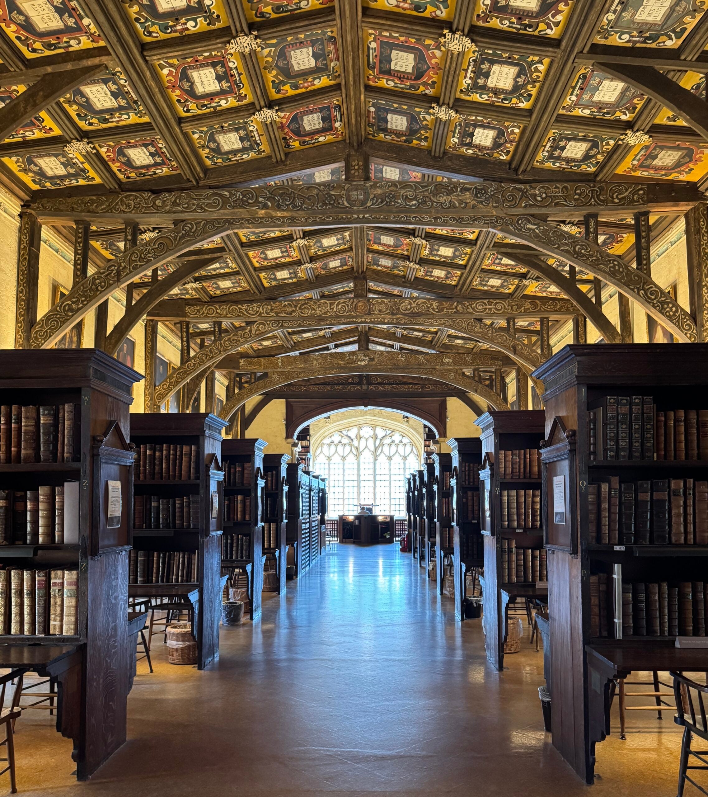 Long view of a library with ornate vaulted ceiling, bookcases on both sides, and a bright window in the distance.