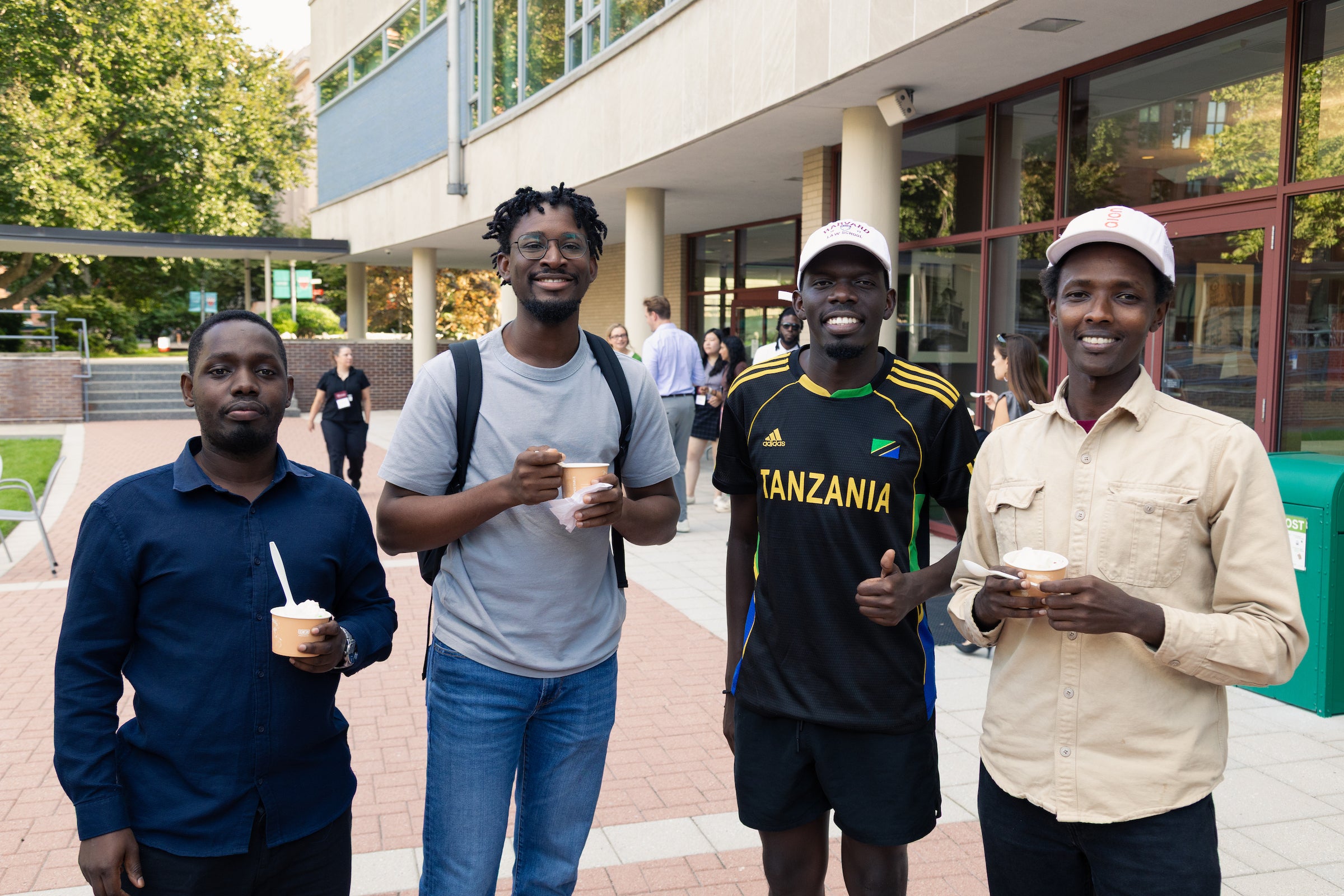 Four students stand outdoors holding cups of ice cream, smiling for the camera in front of a modern building.