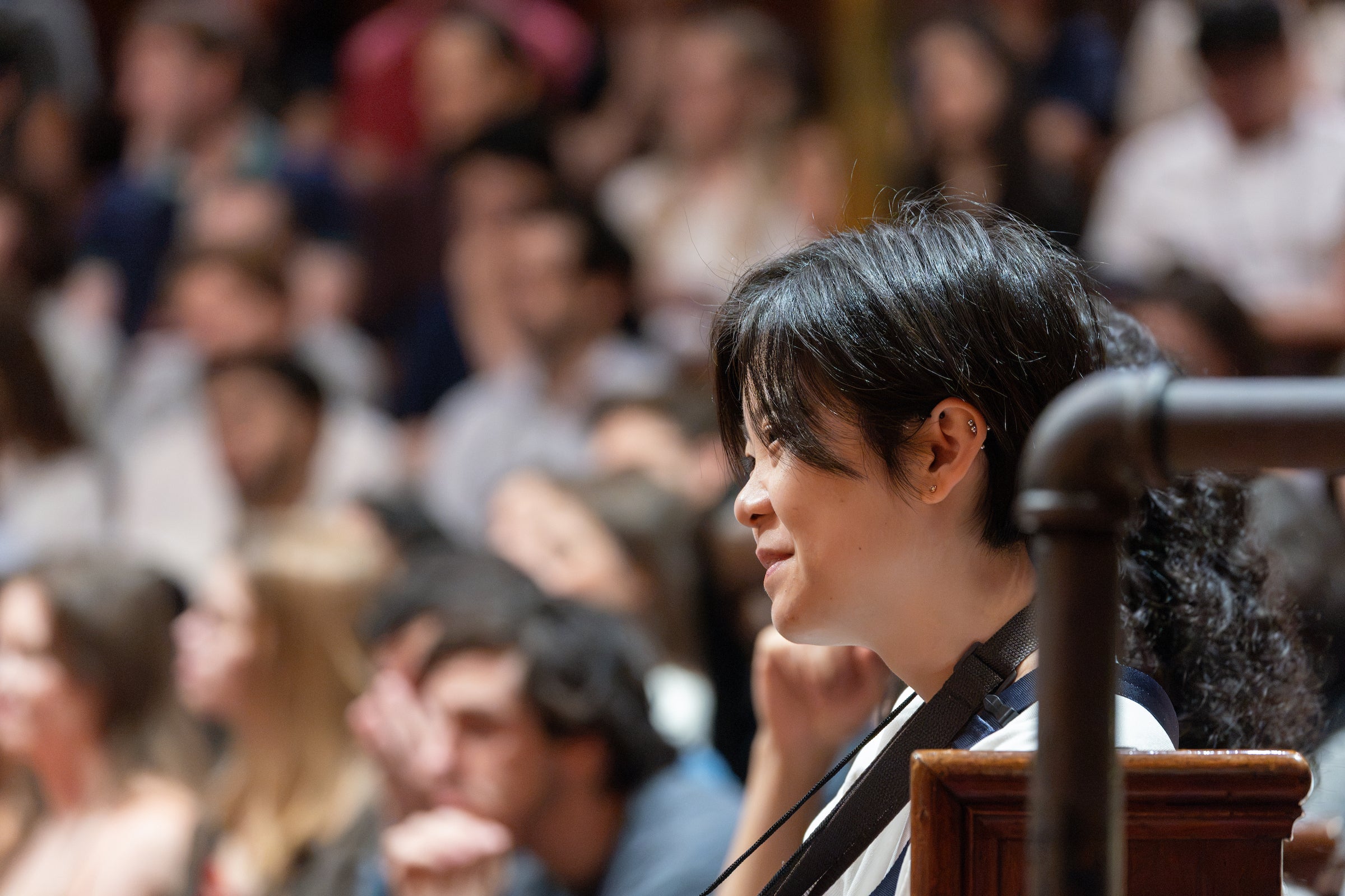 A student sits in an audience, smiling softly while looking toward the stage.