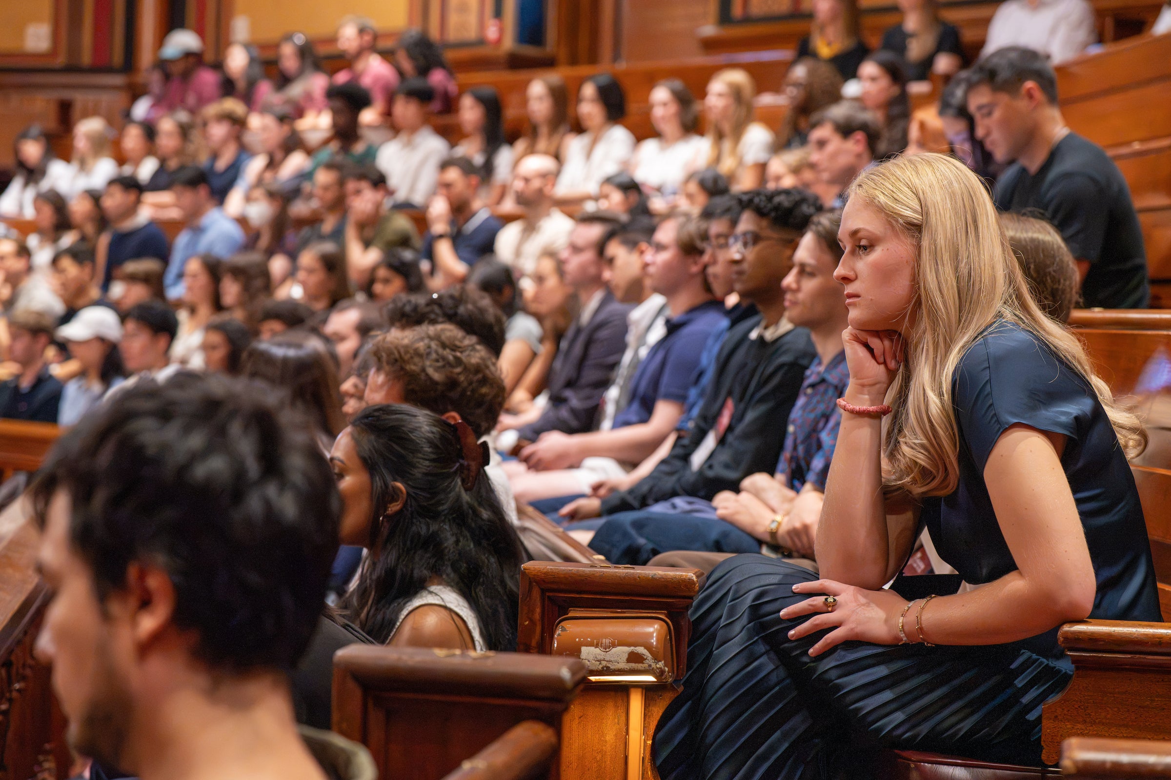 An audience sits in wooden pews, attentively listening in a formal theatre.