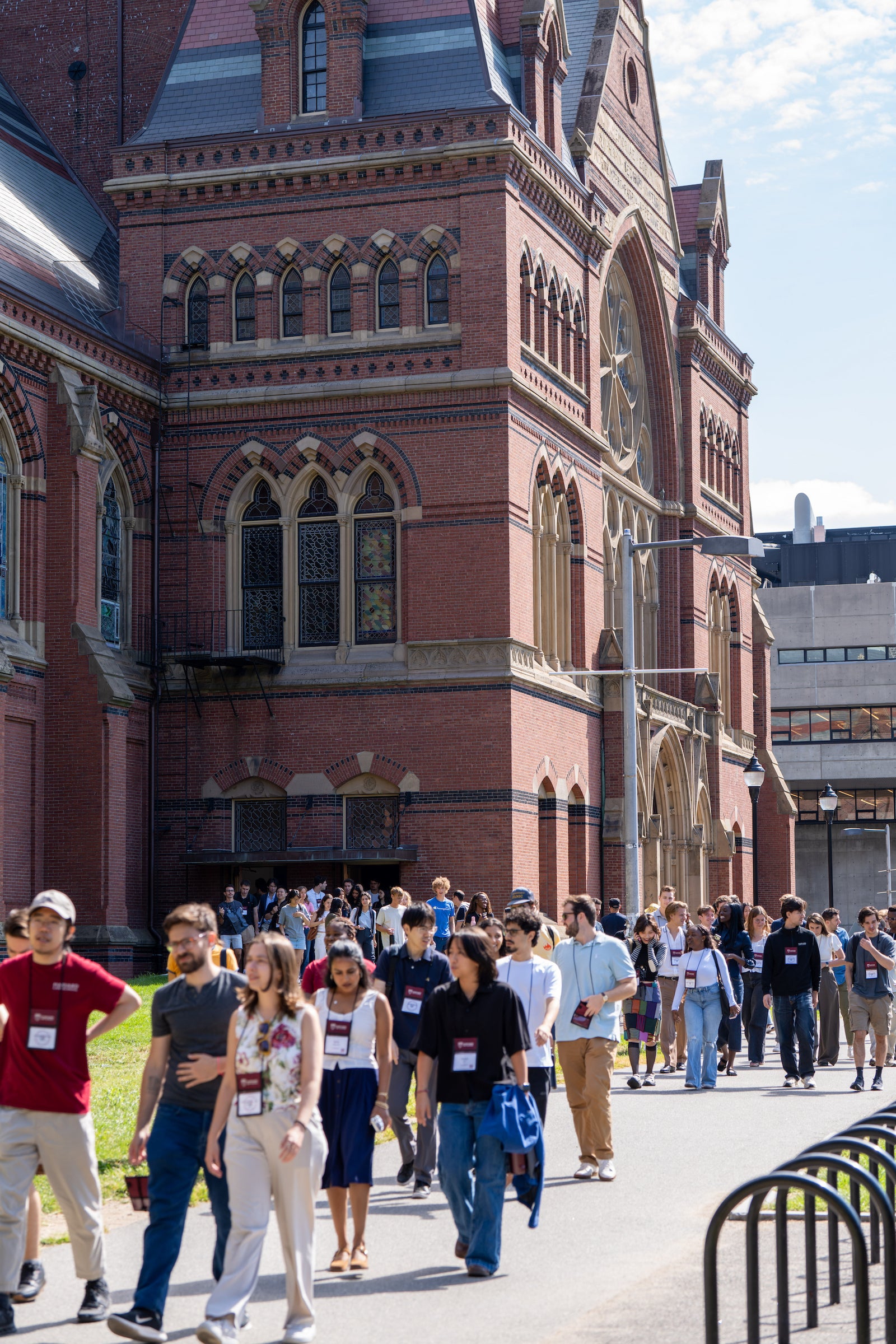 A group of students walk along a path in front of a large, ornate red brick building with arched windows.