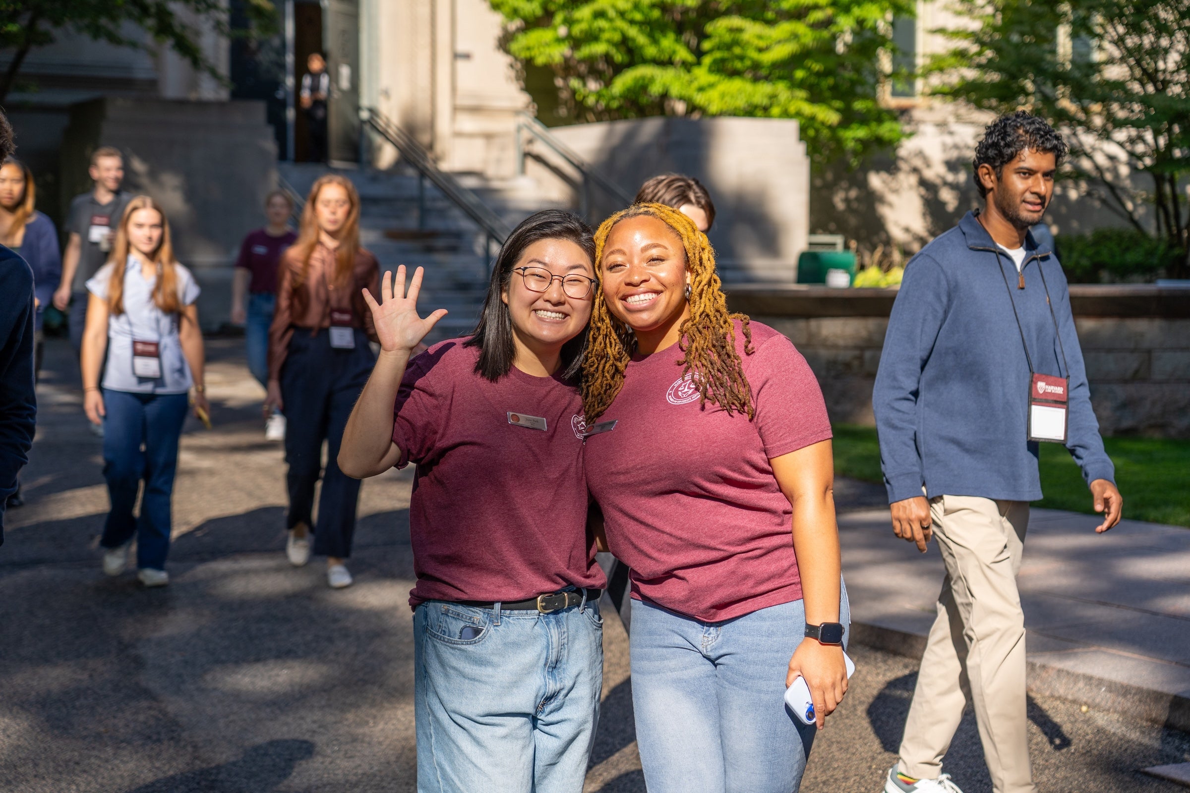 Two individuals in matching maroon shirts smile and wave while walking outdoors with others in the background.