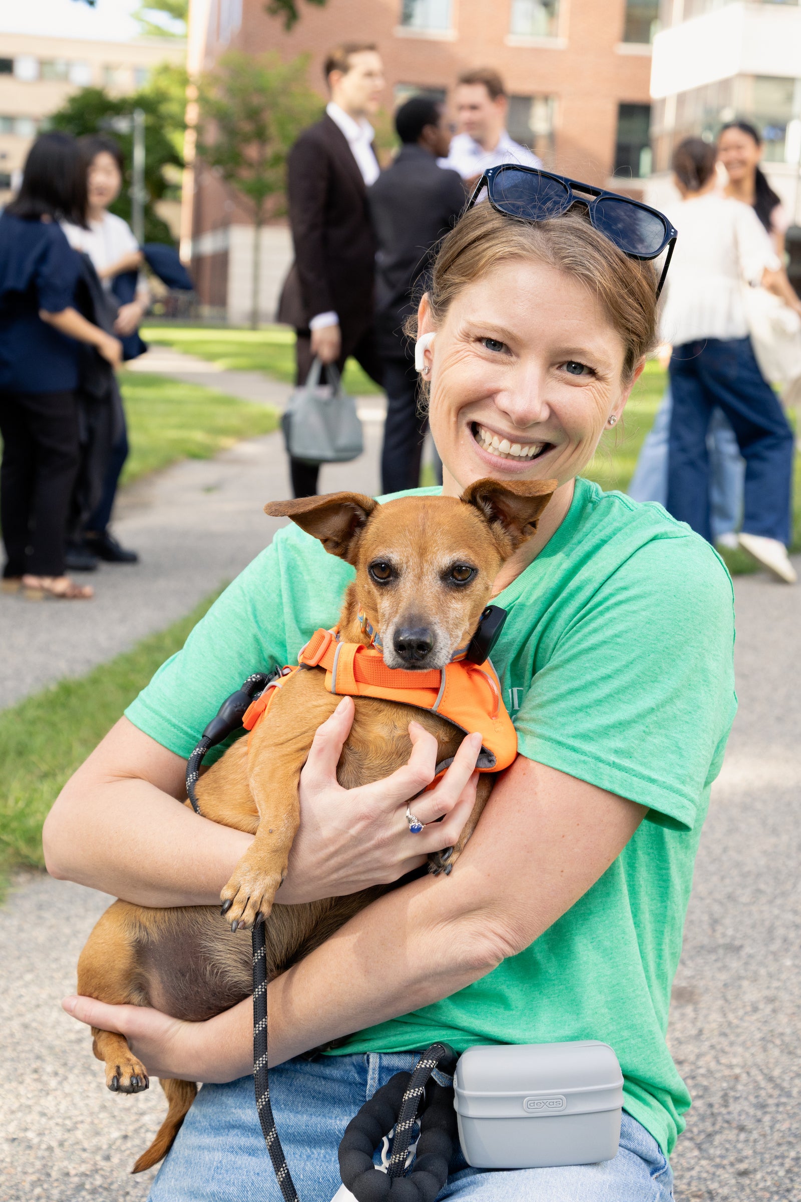 A person in a green shirt smiles while holding a small brown dog wearing an orange harness at an outdoor event.
