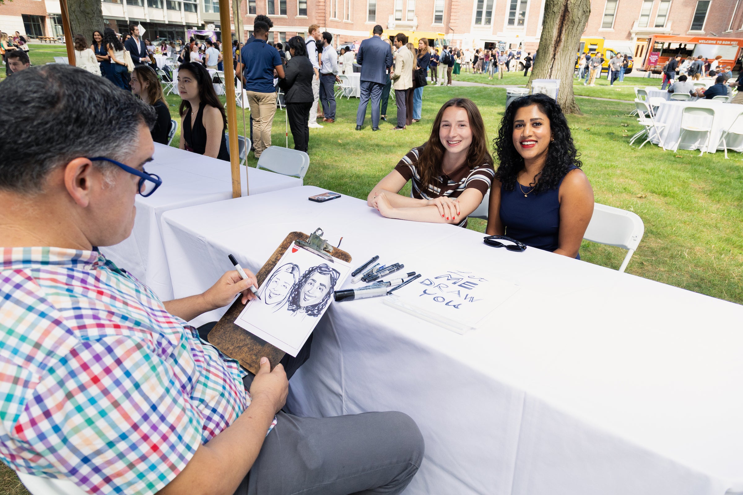 Two students sit at a table and smile for the camera while getting their caricatures drawn.