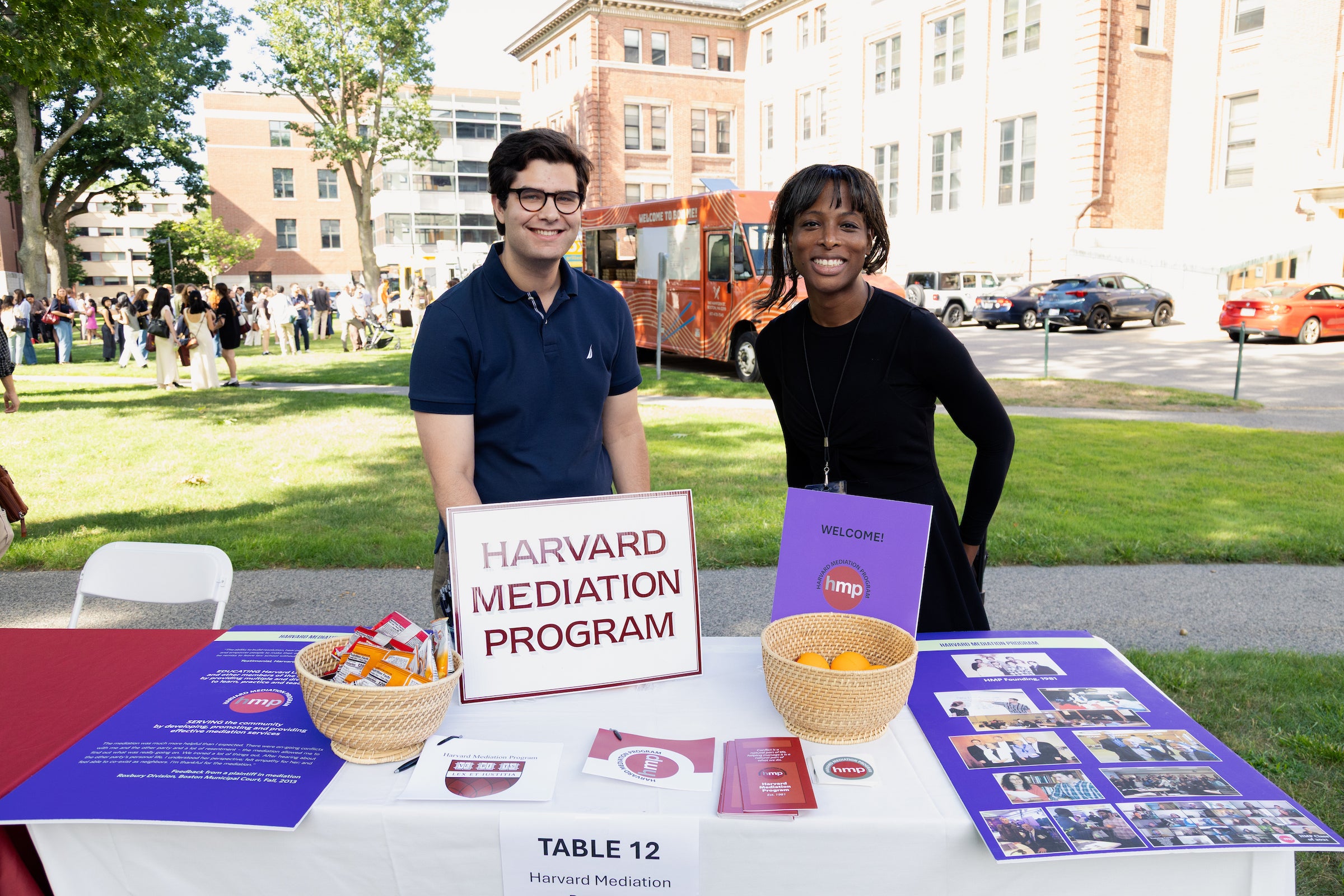 Two individuals smile for the camera while standing behind a table with information for the Harvard Mediation Program.