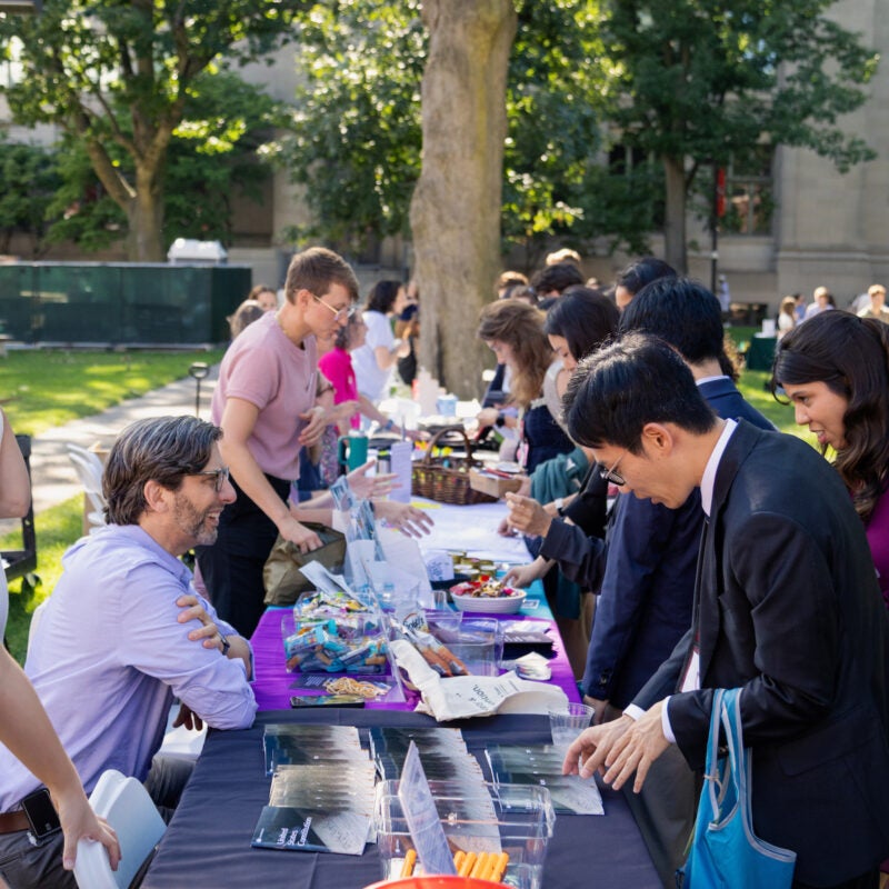 People gather outdoors at a fair around a long table laden with snacks and items, engaging in conversation under the shade of large trees.