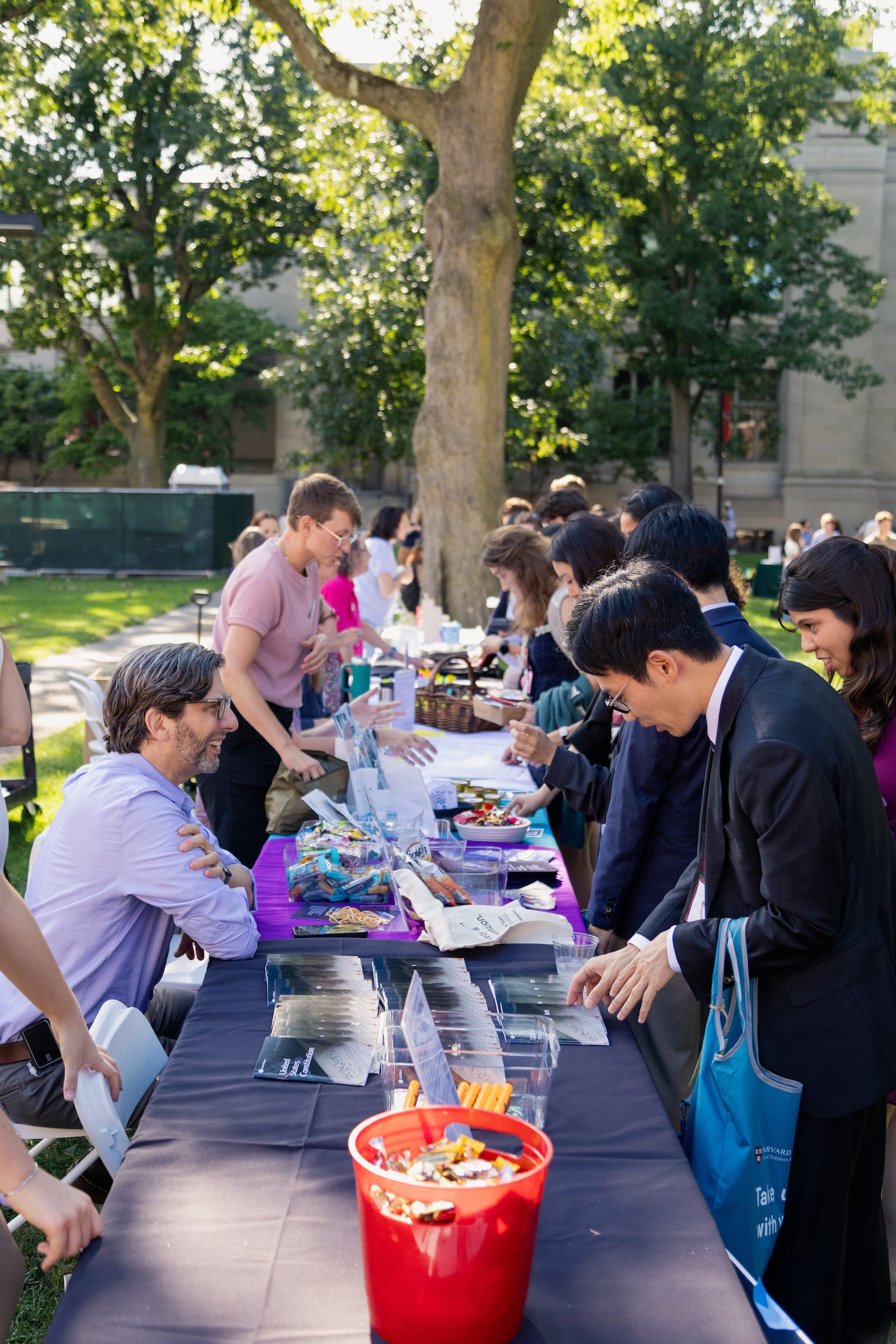 People gather outdoors at a fair around a long table laden with snacks and items, engaging in conversation under the shade of large trees.