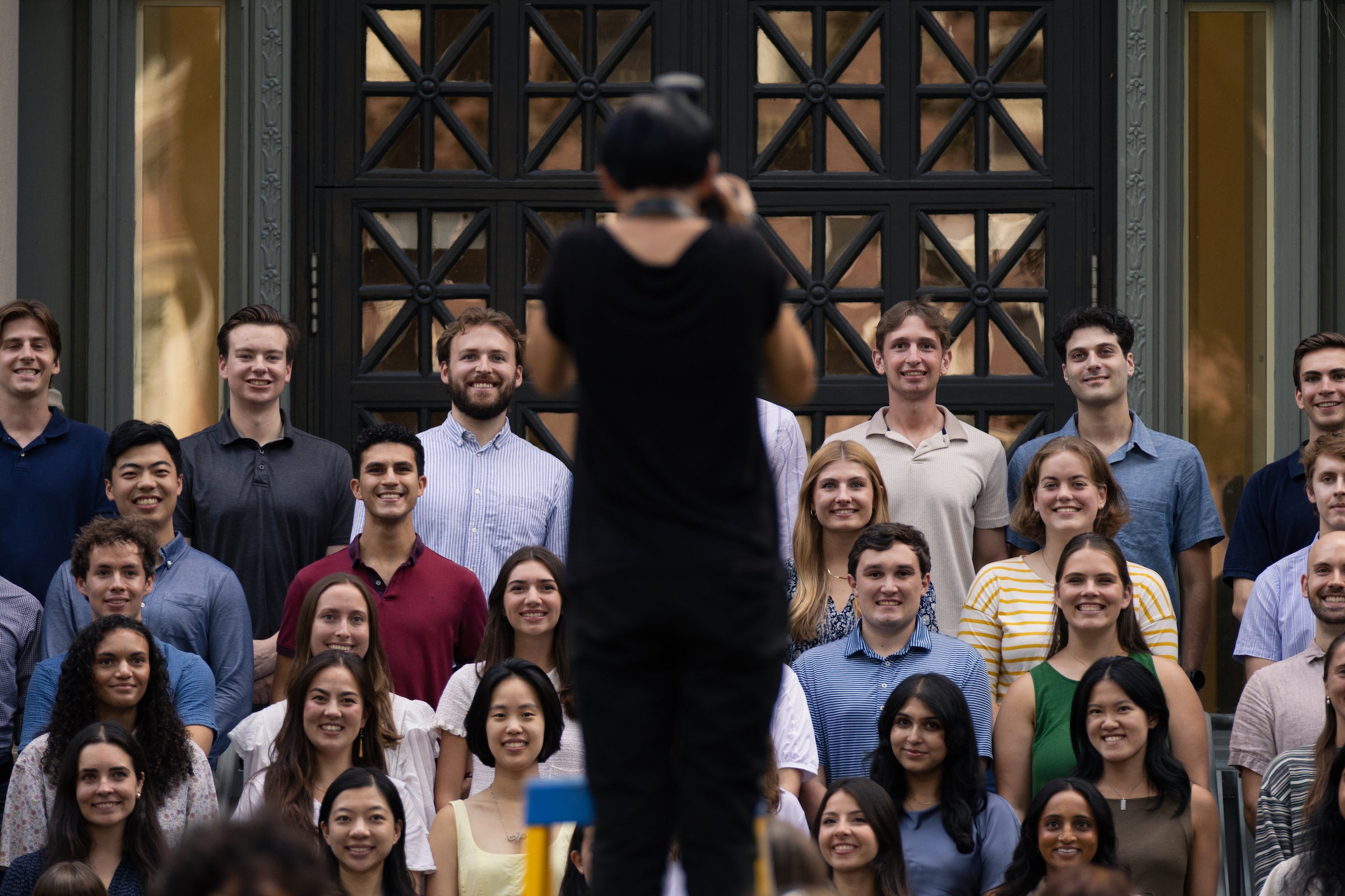 A photographer faces a large group of smiling individuals gathered in front of an ornate building entrance.